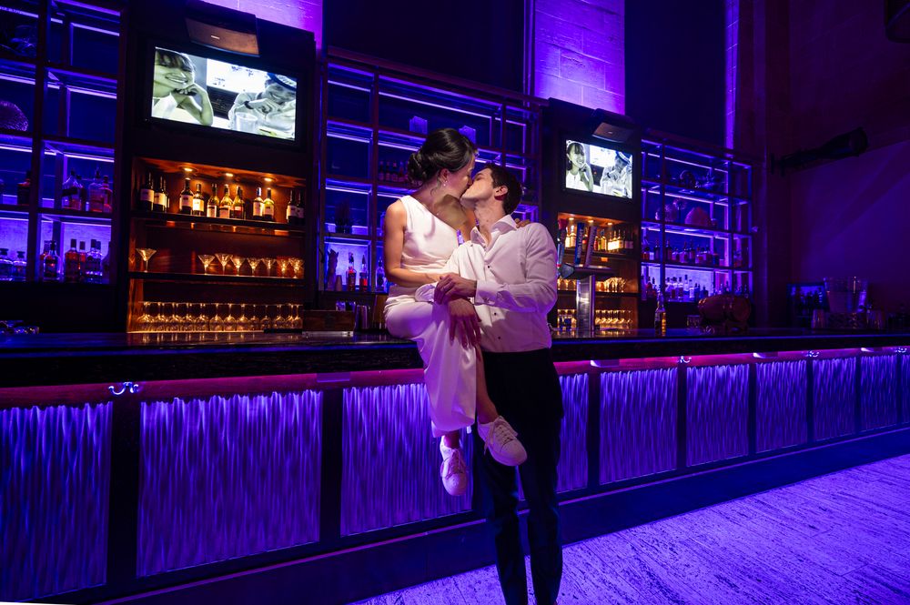 A couple dances together in front of an illuminated bar with purple lighting and TV screens.