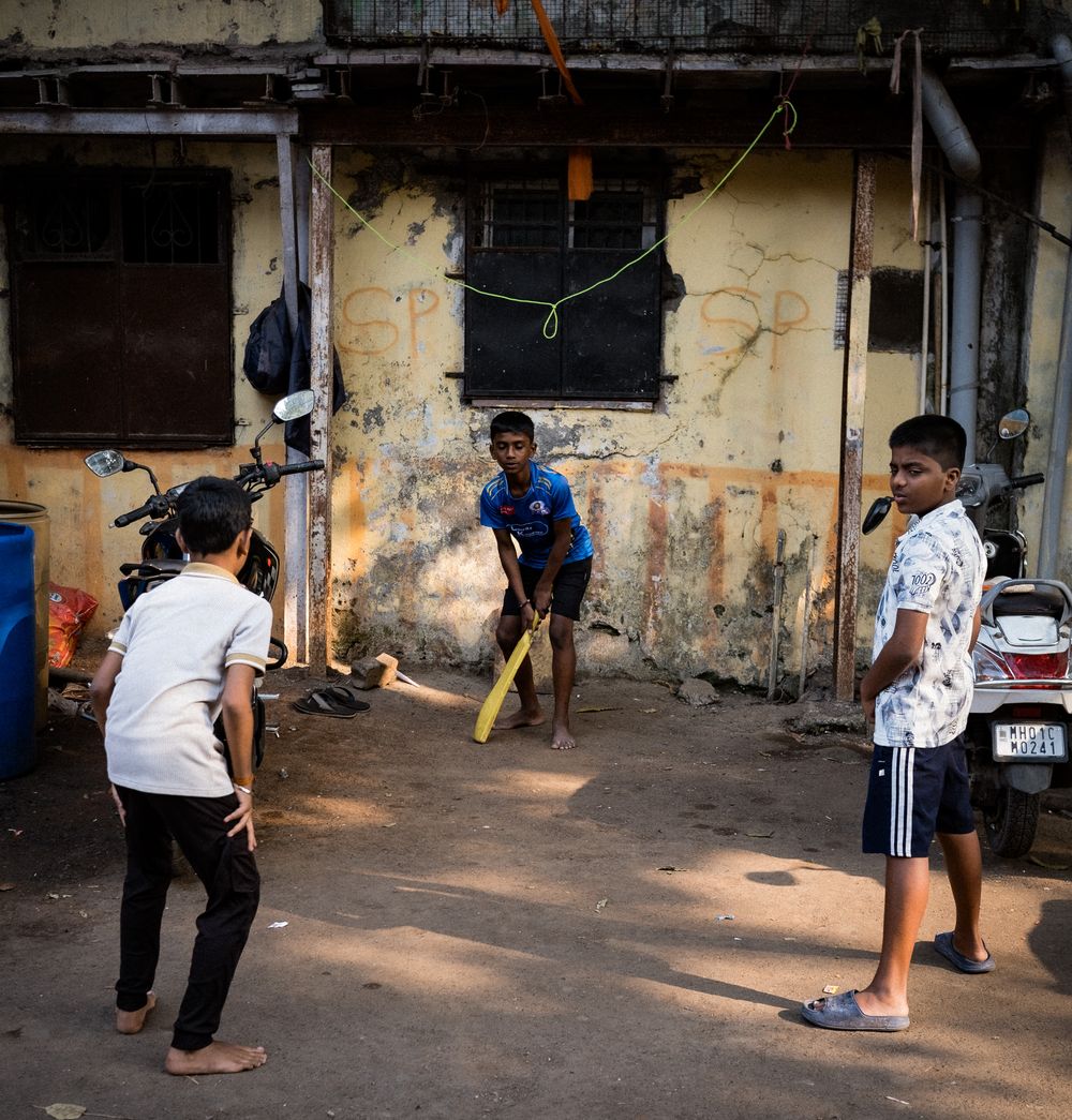 Three boys play cricket in a narrow urban alleyway with weathered yellow walls and a parked motorcycle.