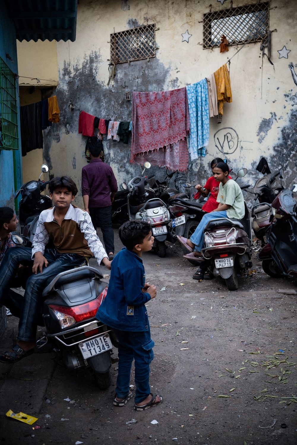 Children and teenagers gather in a crowded urban courtyard with parked scooters and laundry hanging on walls.
