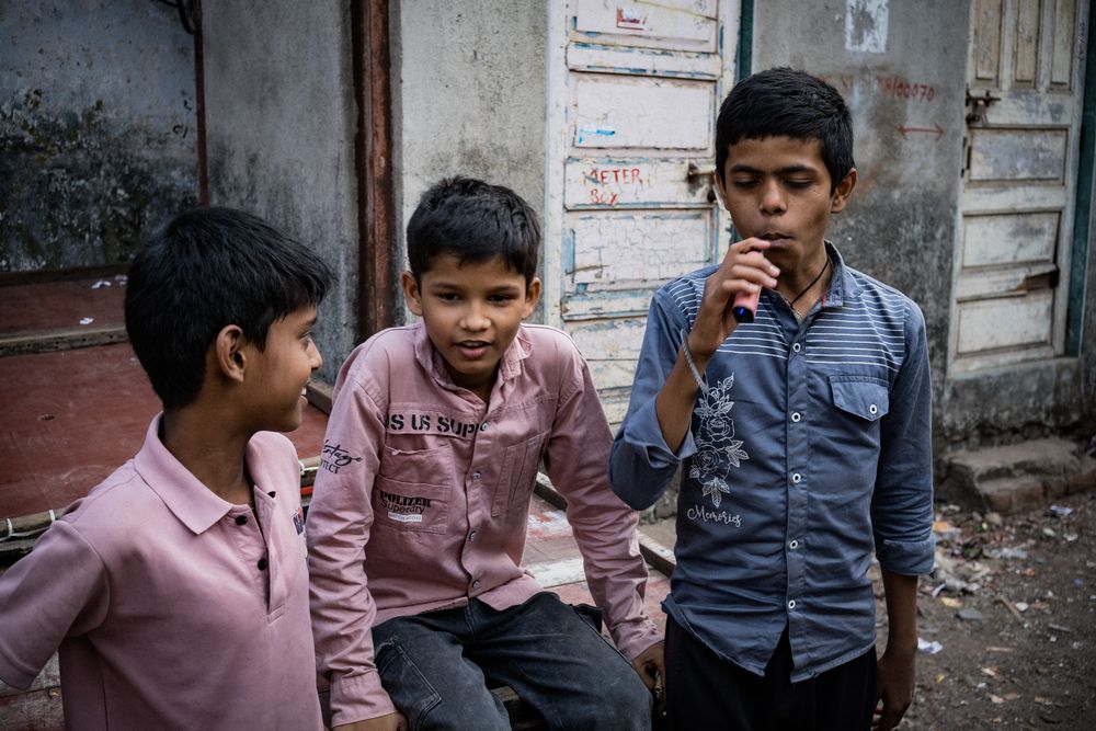 Three Indian boys sitting outside a weathered building, one eating a snack in an urban neighborhood.