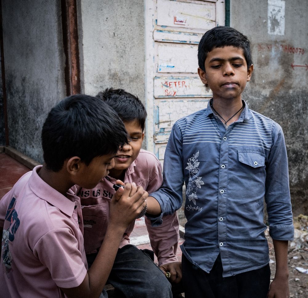 Three Indian boys gathered outside a worn building wall, one child eating while others watch closely.