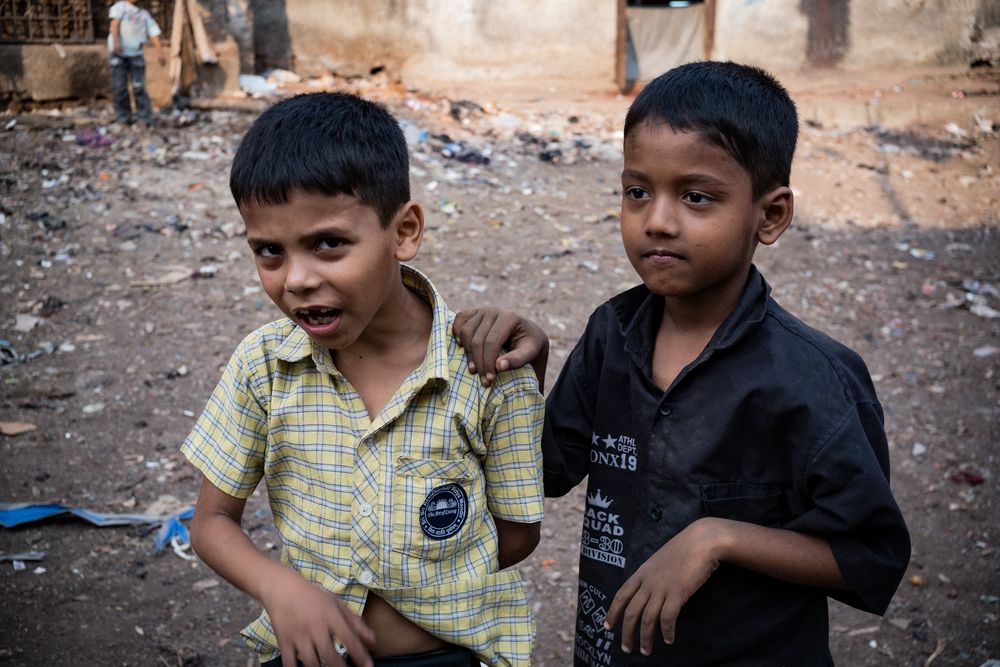 Two young Indian boys standing together in a dusty outdoor area, one with arm around the other.