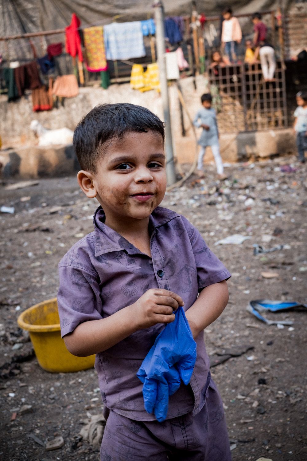 A young Indian toddler in a dirty purple shirt holding a blue plastic bag in a slum area.