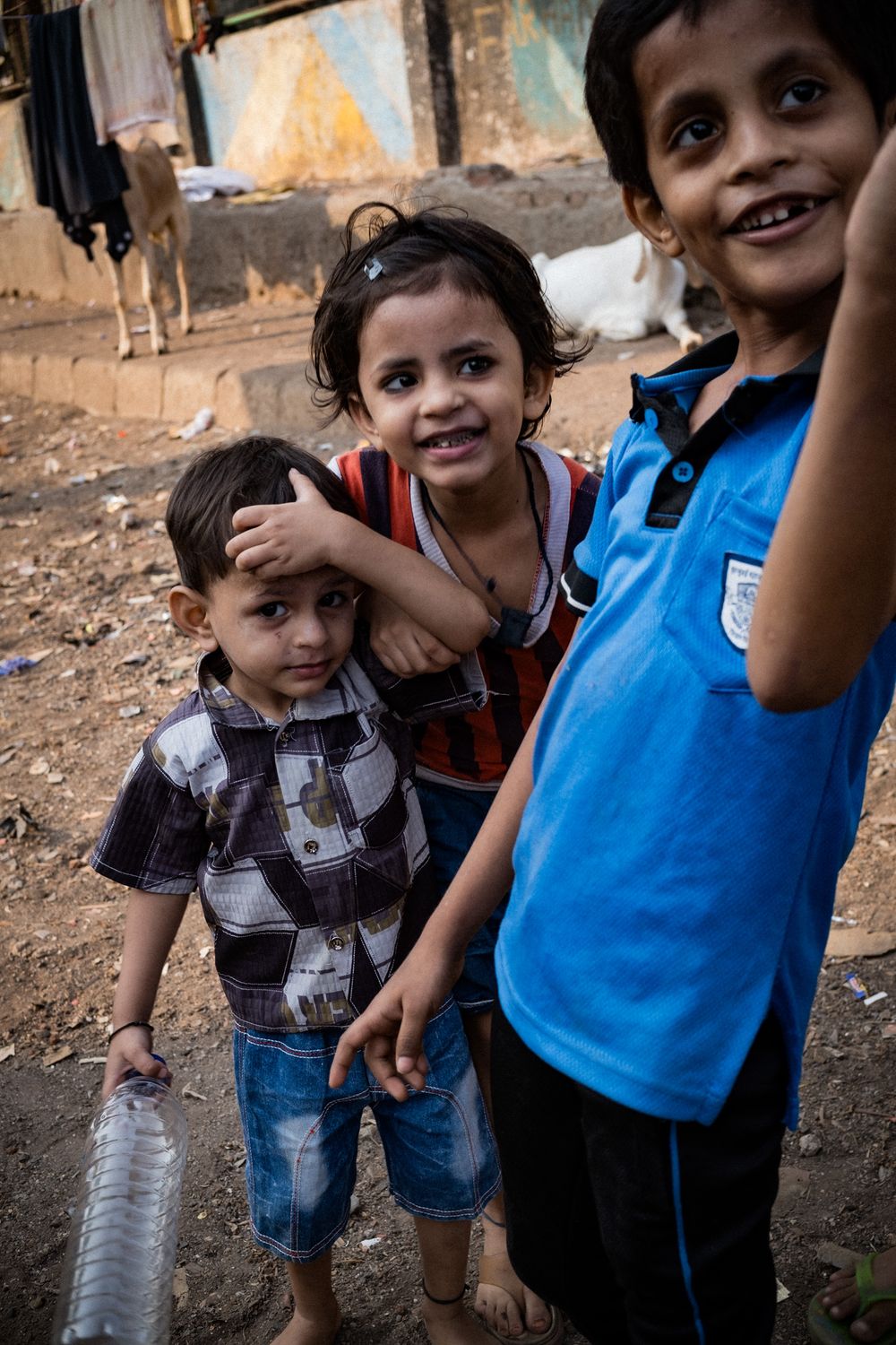 Three young Indian children posing together outdoors in a rustic neighborhood setting with a cow nearby.
