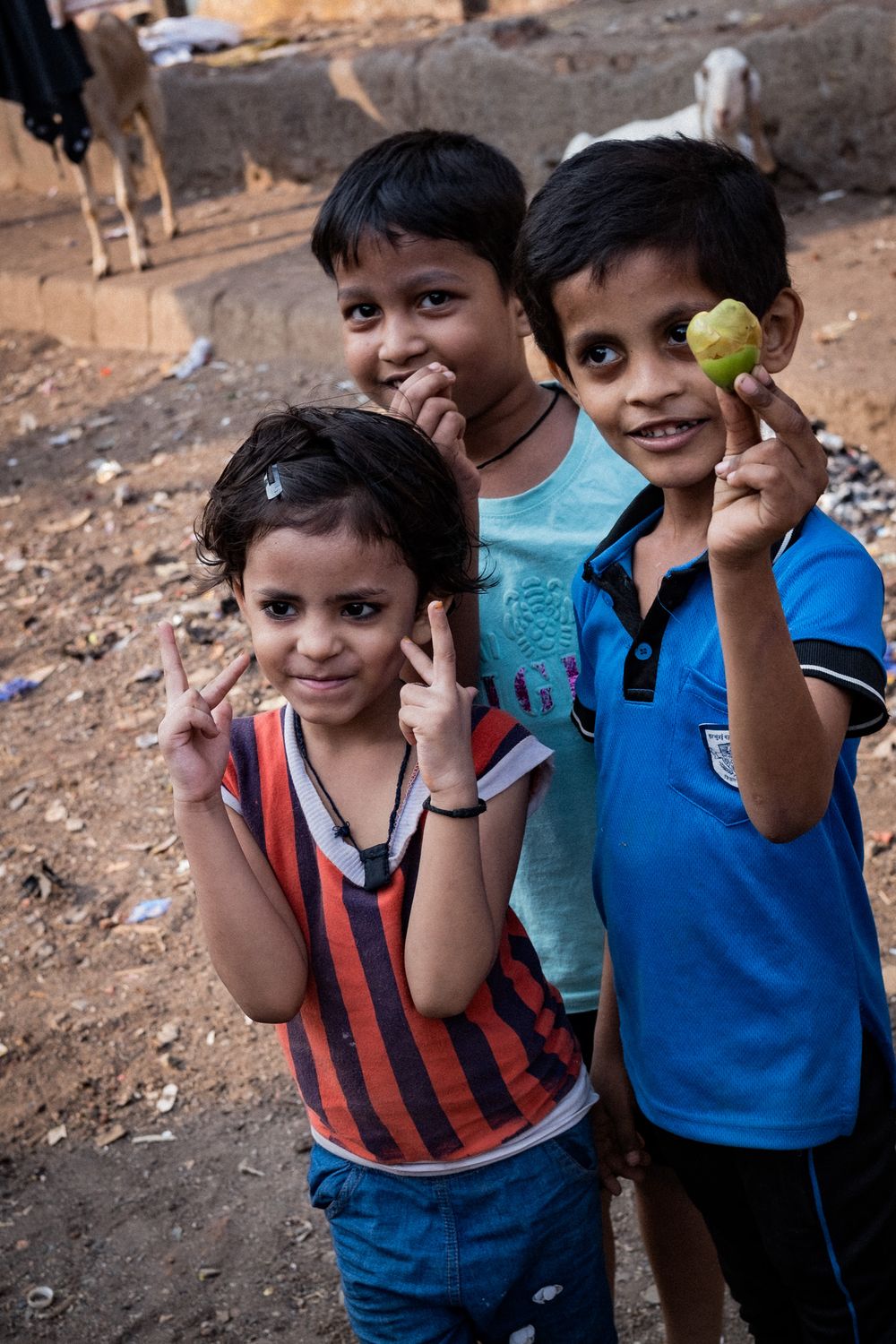 Three smiling Indian children posing outdoors, one flashing peace signs and another holding a small fruit.