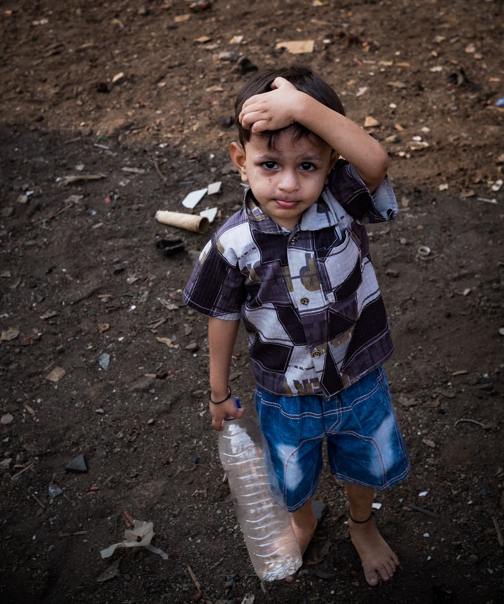 A young Indian boy in a plaid shirt holding a plastic water bottle while standing on bare ground.