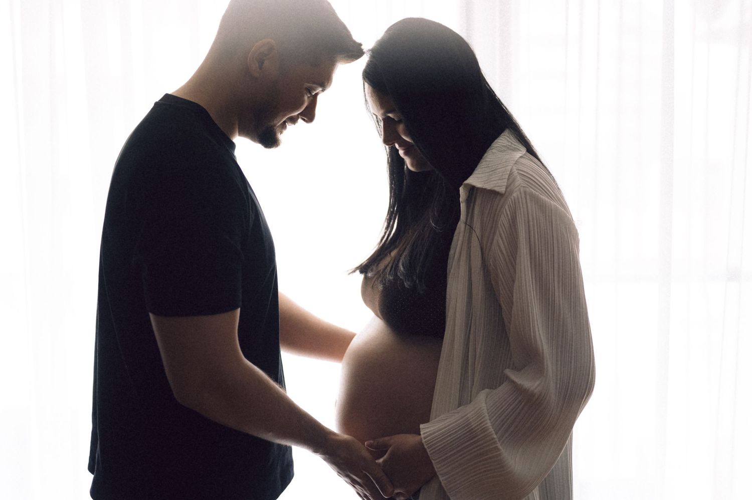 A couple stands in silhouette against a bright background during an intimate moment together.