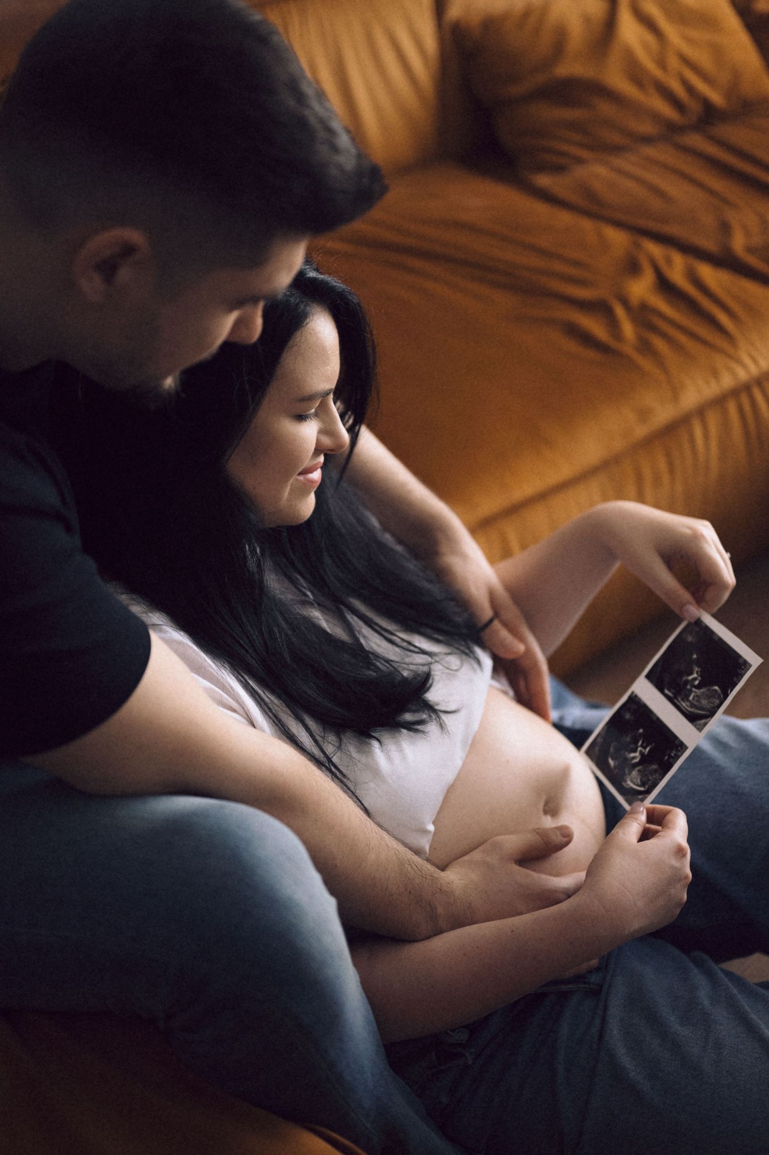 A couple looks at an ultrasound image on a mobile phone while sitting together on a brown leather couch.