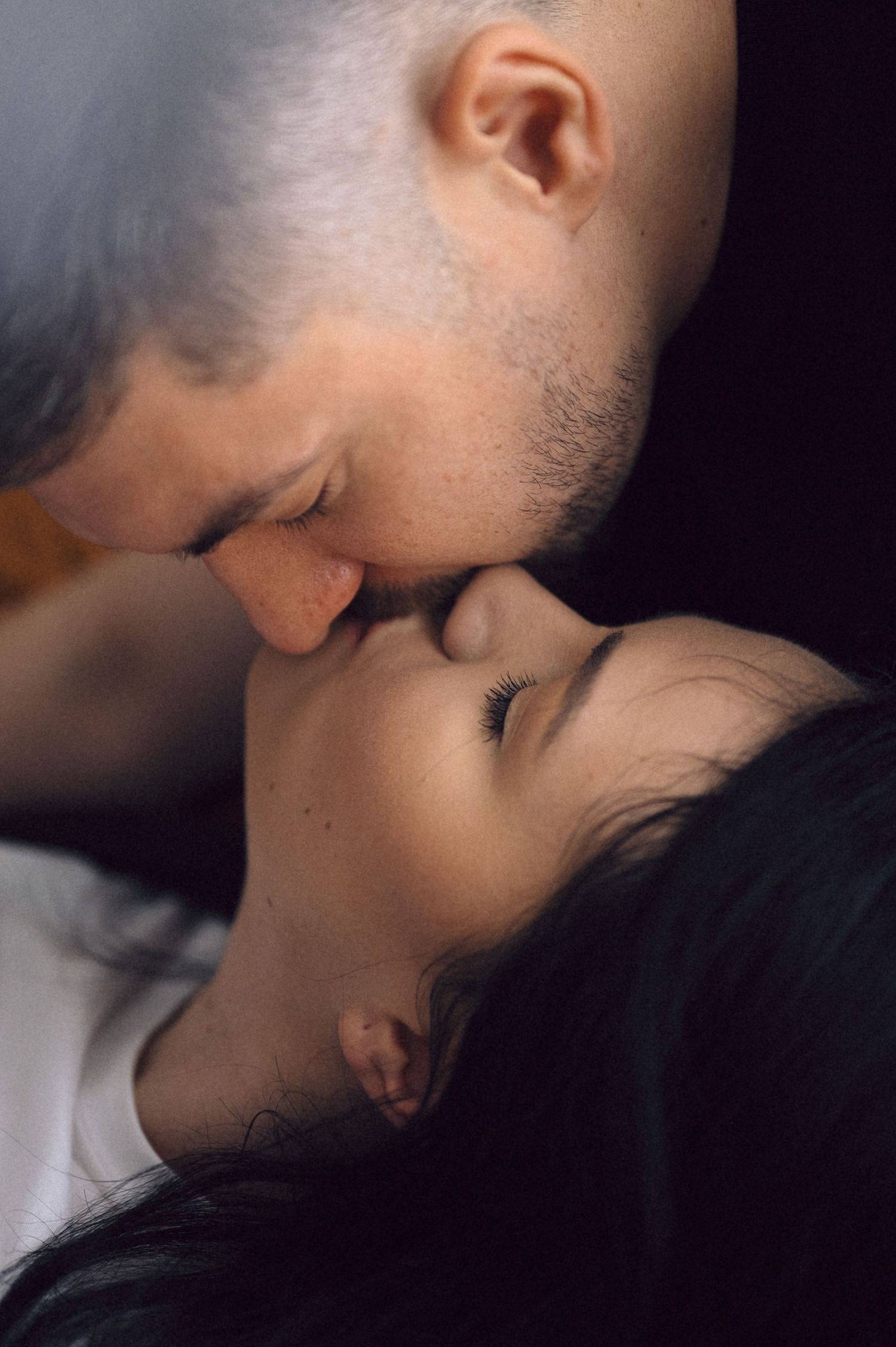 An intimate close-up portrait of a tender romantic moment between two people against a dark background.