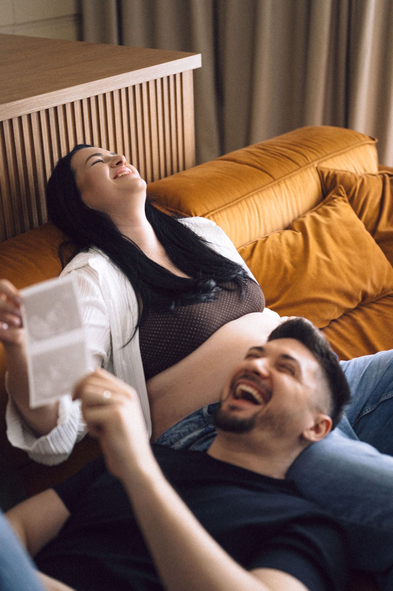Two people lying on a couch laugh together in a warm, sunlit room.