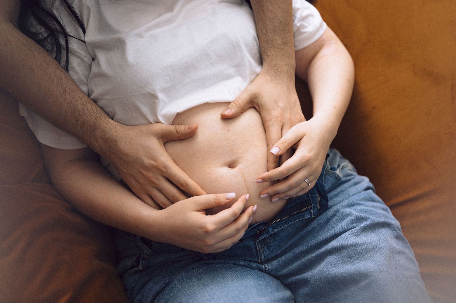 Close-up view of hands folded in lap wearing jeans and white shirt against brown leather background.