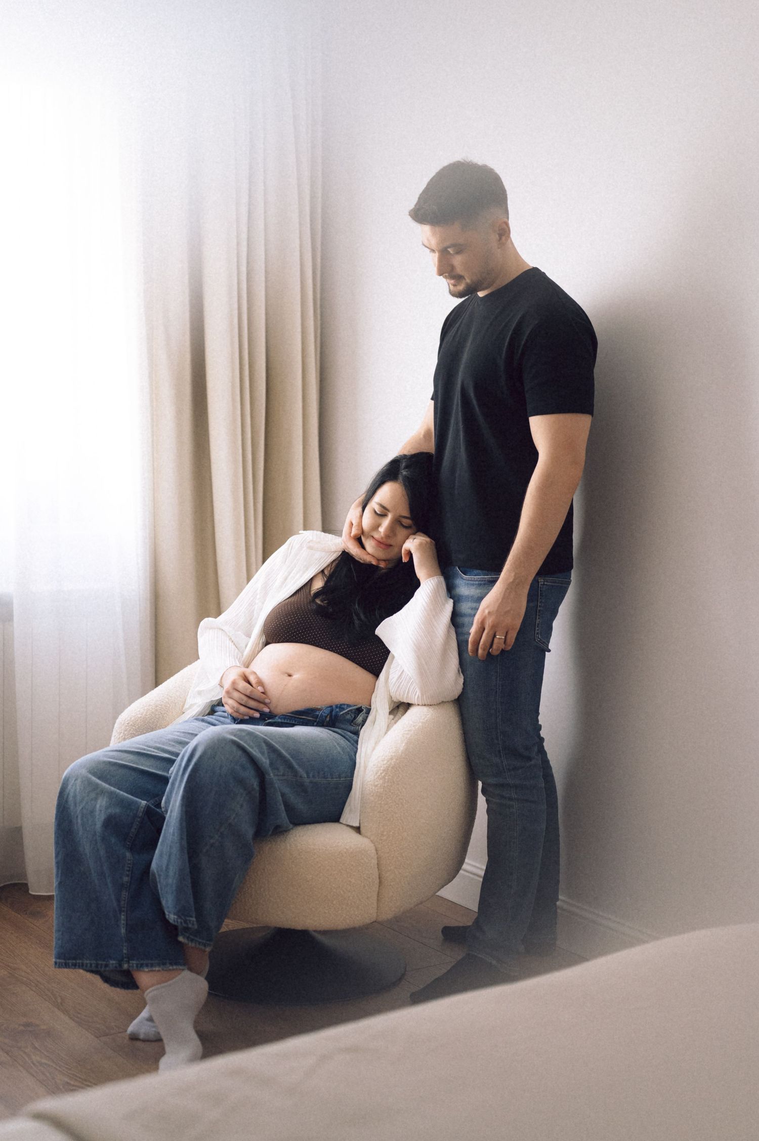 Couple sharing intimate moment in living room as pregnant woman rests on beige armchair near curtained window.