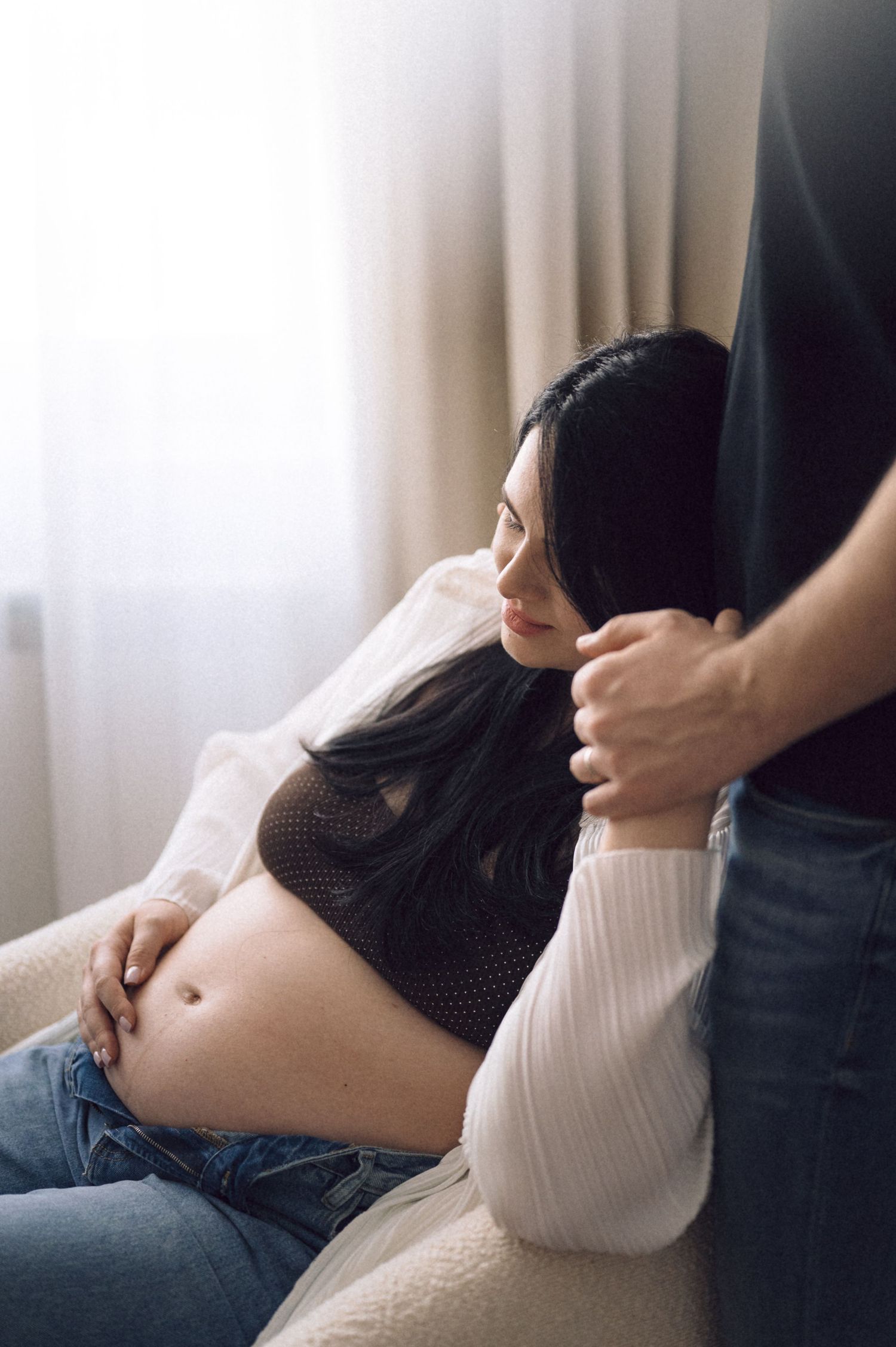 A series of intimate maternity photos showing a pregnant woman in a white top reclining on a couch in natural light.