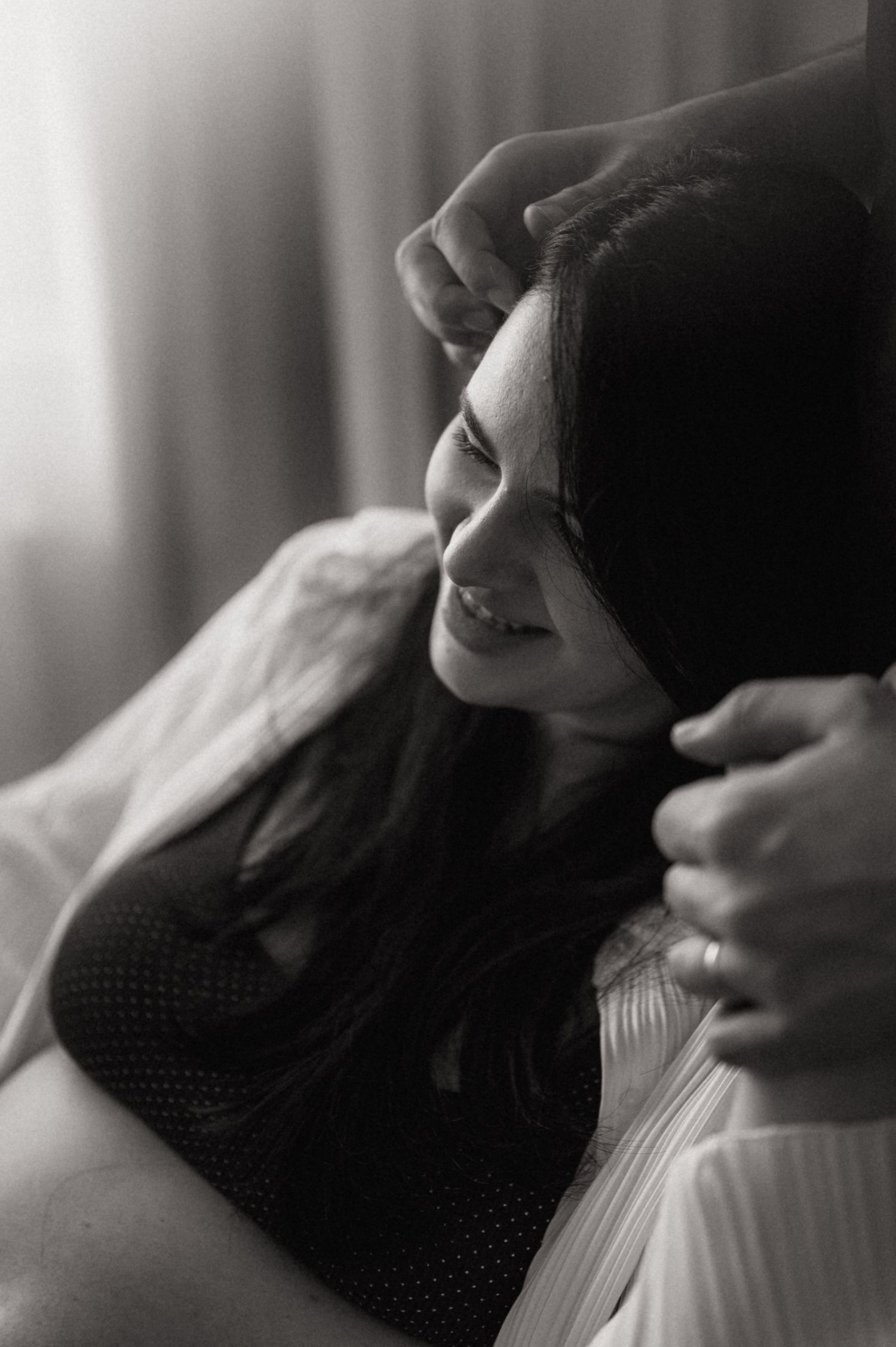 A black and white portrait photograph capturing a candid moment of joy and laughter in soft window light.