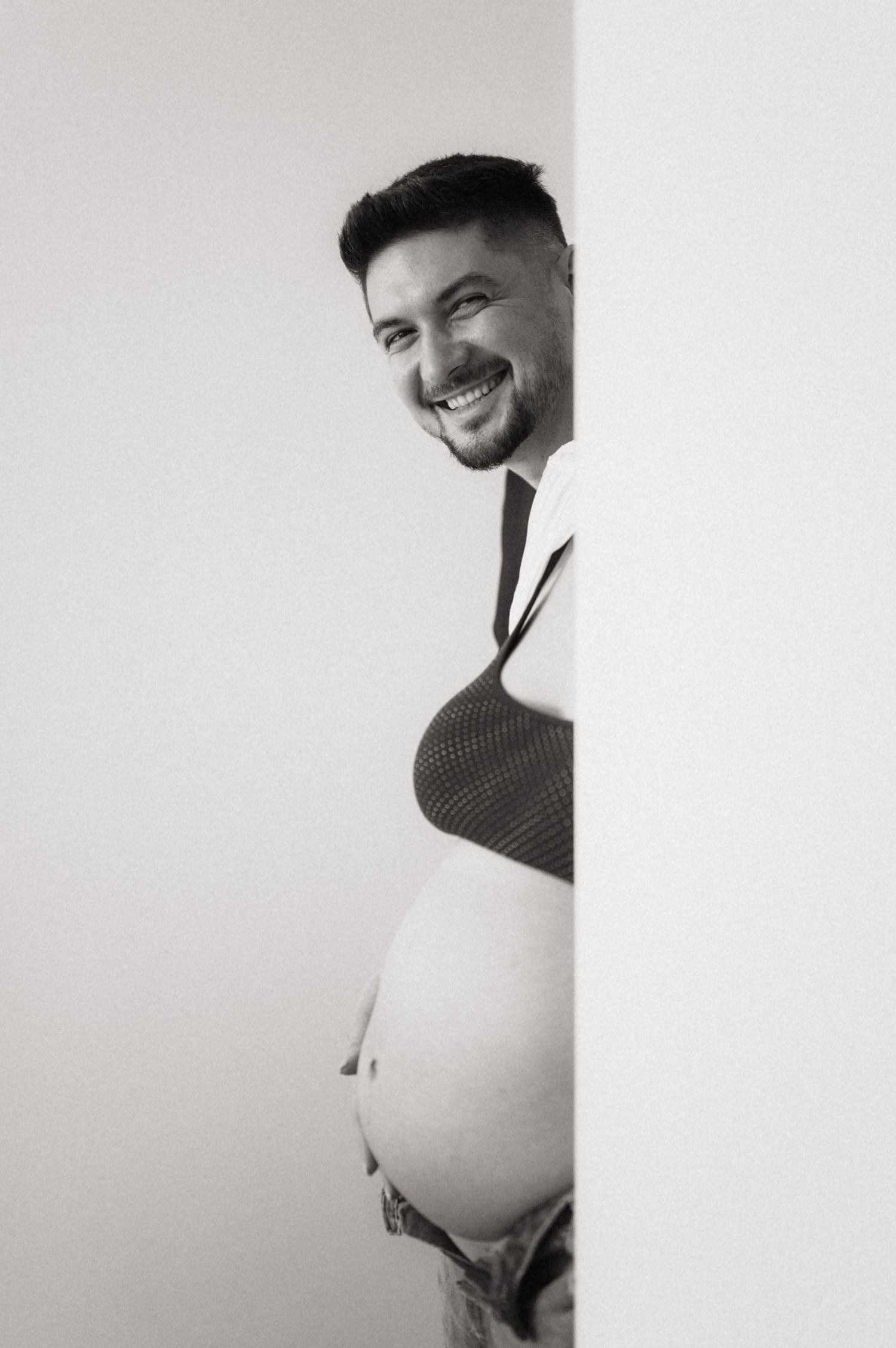 Black and white portrait photo showing someone peeking around a wall corner with a bright smile.