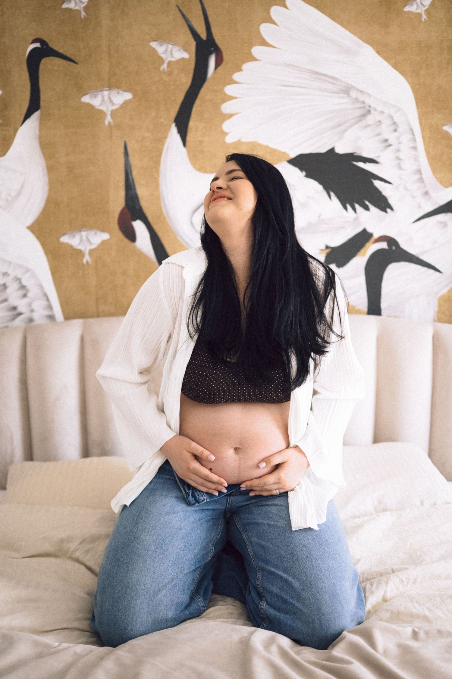 A person in jeans sits on a bed with white paper birds decorating the wall behind them.