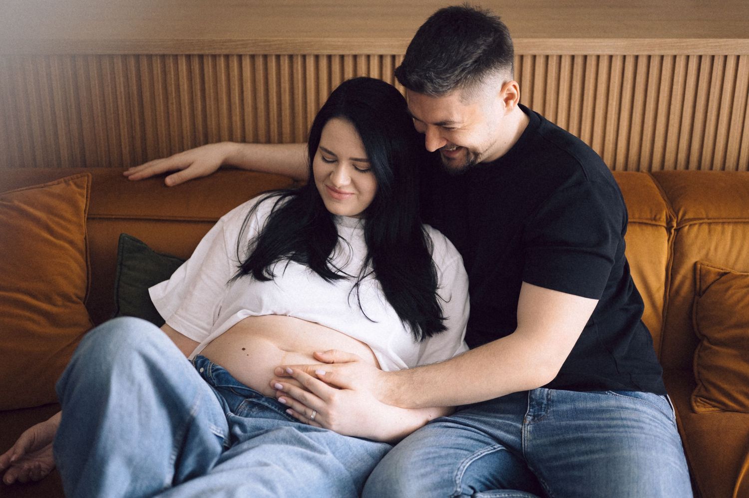 Couple sharing intimate moment on couch during maternity photoshoot in warm lighting.