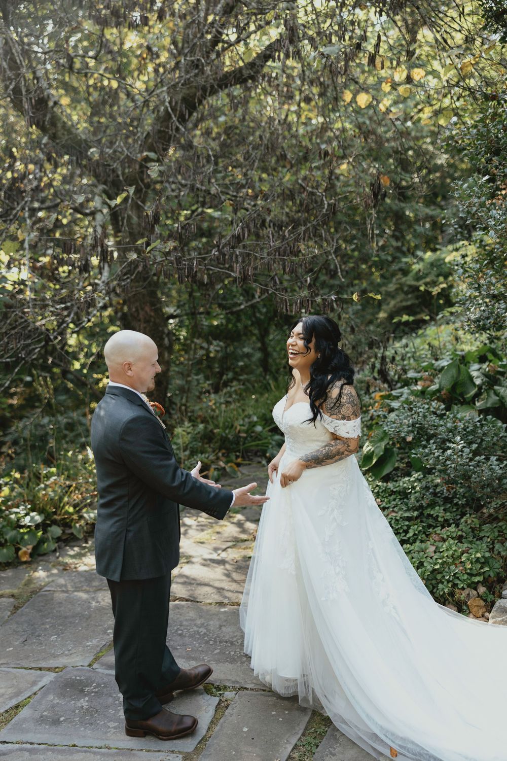 A couple in wedding attire shares a playful moment on stone steps surrounded by lush greenery in a garden setting.