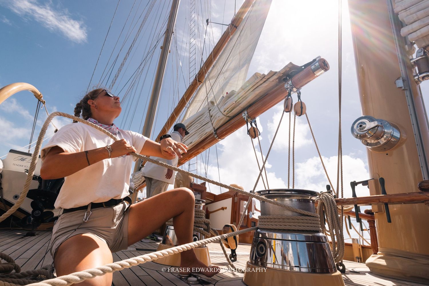 A person adjusts the rigging on a sailboat deck while sunlight streams through the white sails on a clear day.