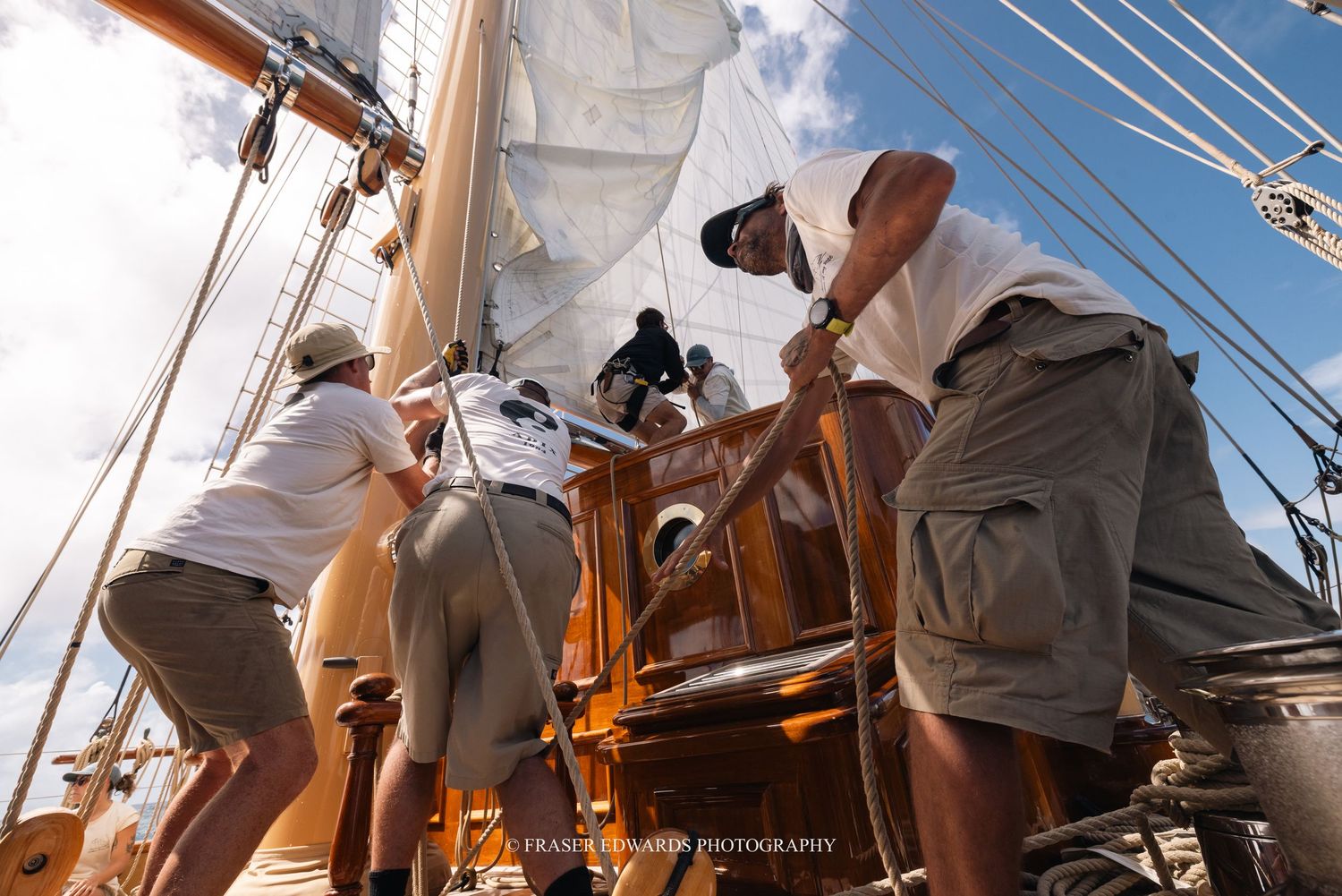 Crew members work together to adjust sails and rigging on a traditional sailing vessel during a sunny day at sea.