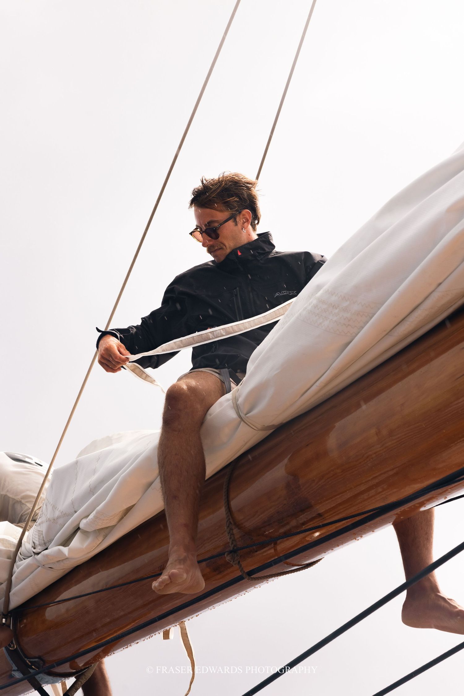 Person adjusts the rigging while perched on the wooden boom of a traditional sailing vessel.