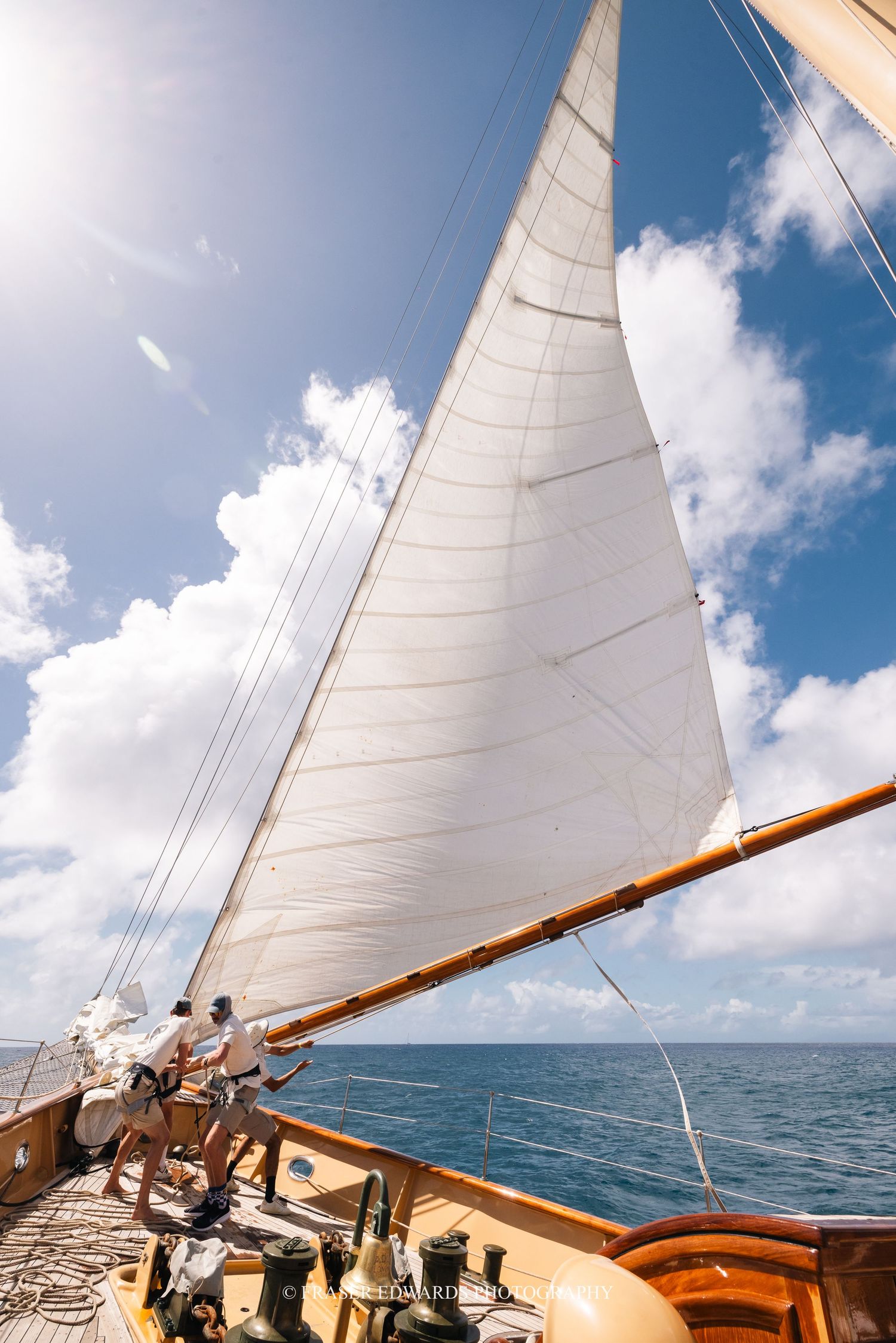 White sails billow against a blue sky with fluffy clouds as seen from the deck of a wooden sailboat.