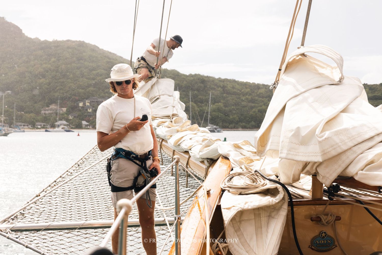 A sailor adjusts rigging and sails on the deck of a yacht with mountains and water in the background.