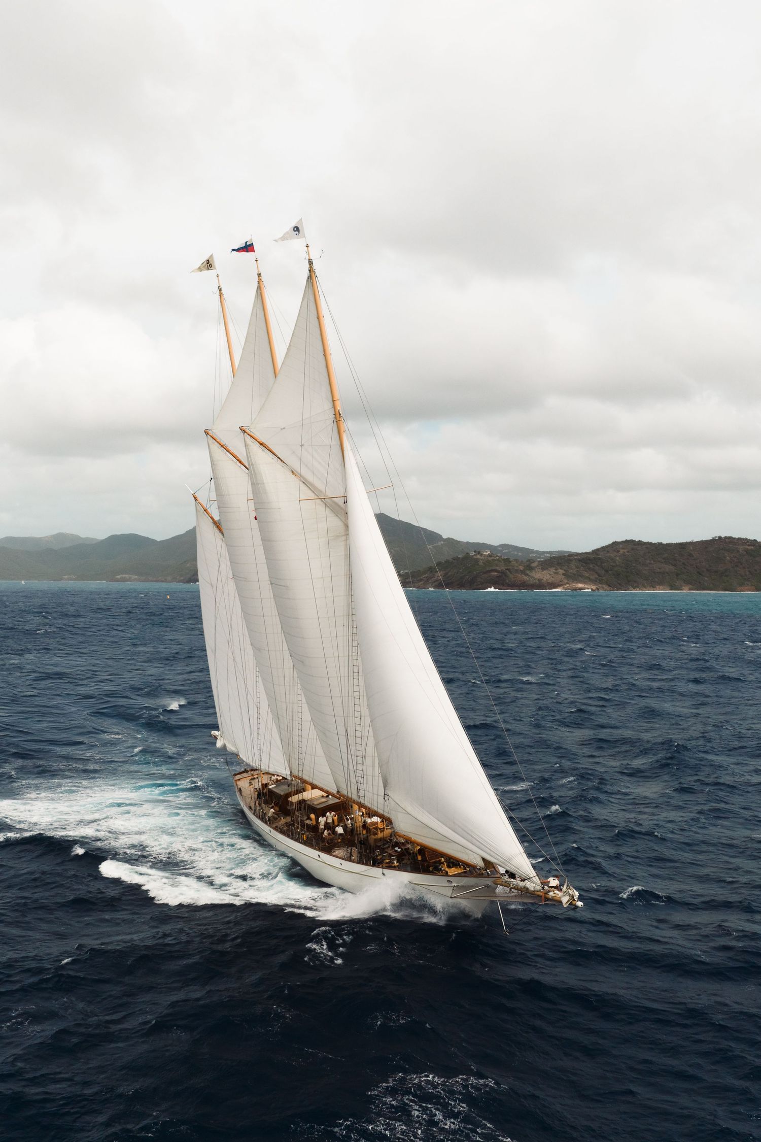 A majestic three-masted sailing ship with white sails glides across blue ocean waters near a mountainous coastline.