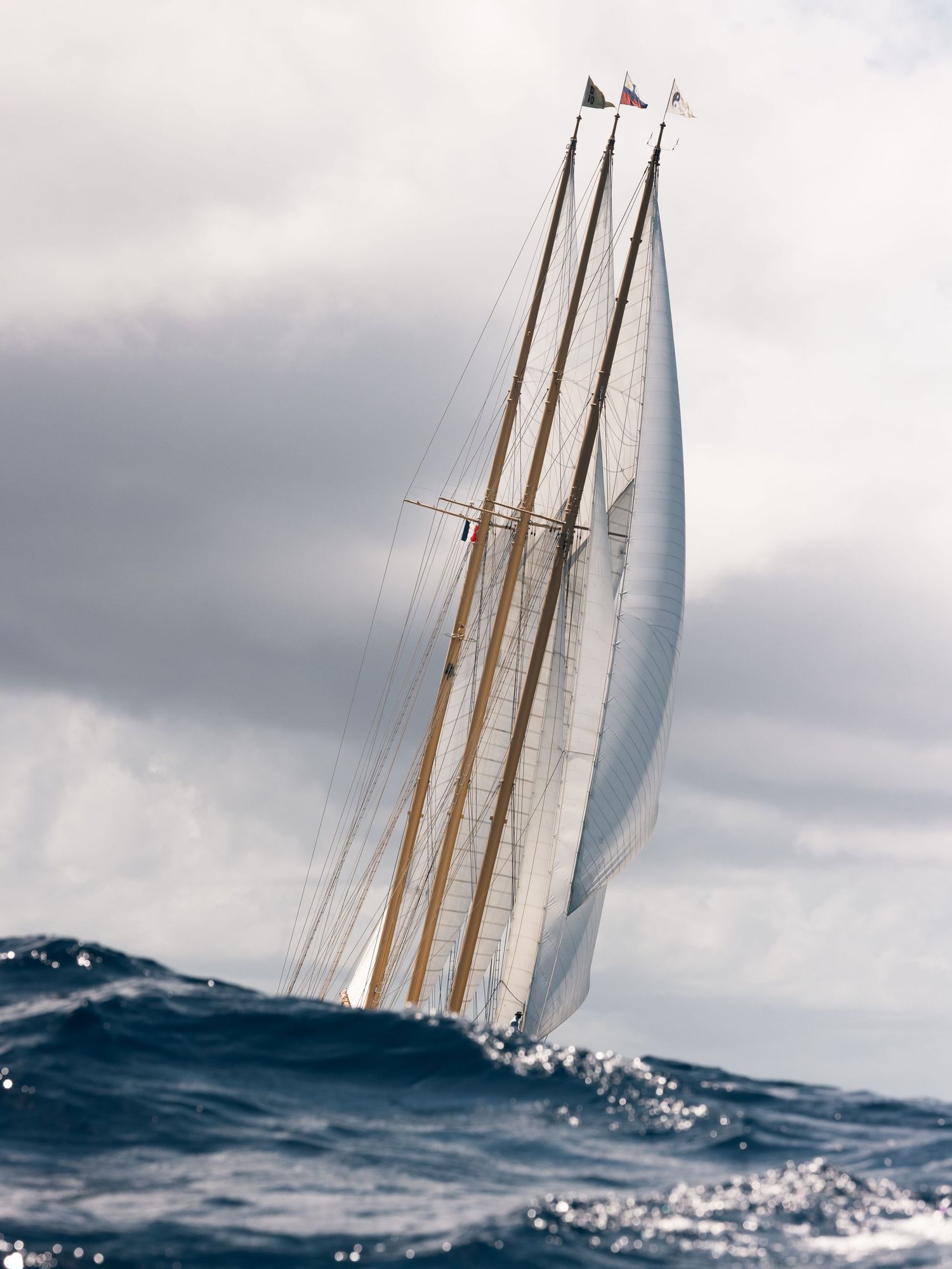 A majestic sailing yacht with three tall masts glides through choppy ocean waters under cloudy skies.