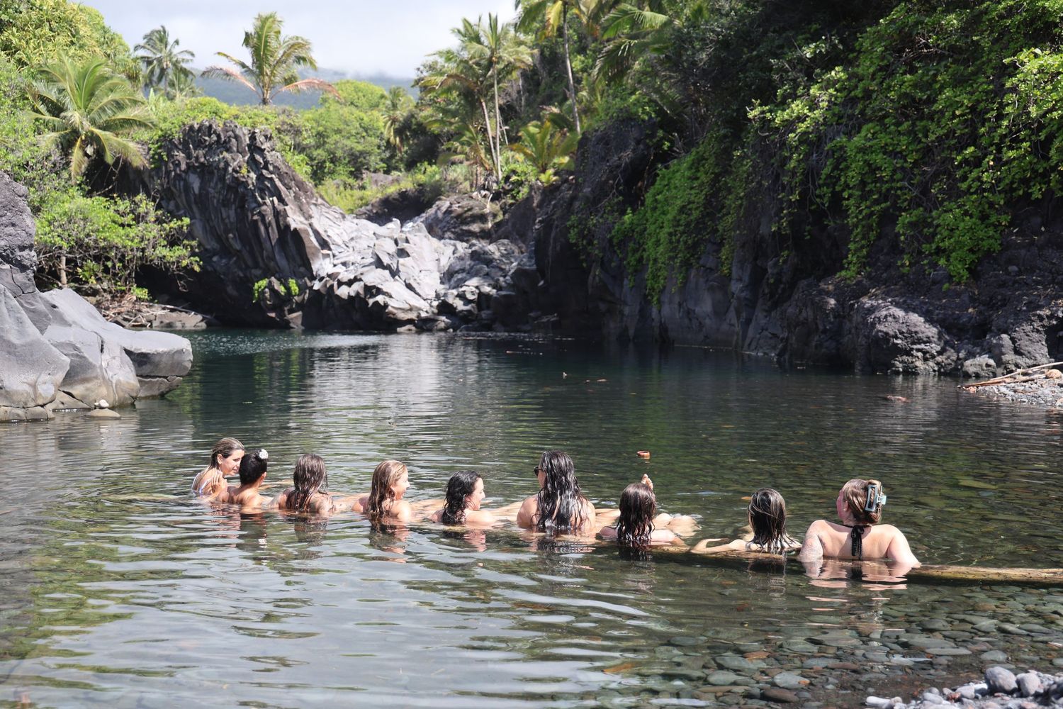 A group of swimmers enjoy a scenic swimming hole surrounded by volcanic rocks and palm trees in a tropical setting.