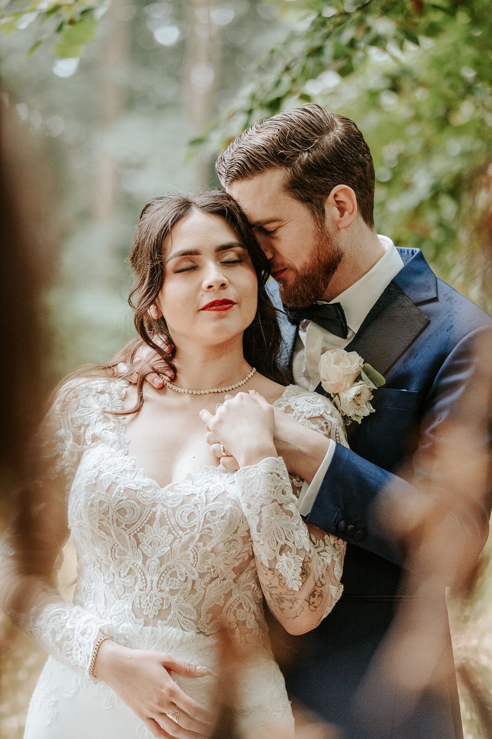 Bride and groom share a romantic embrace in a forest setting on a rainy day, bride in lace gown, groom in navy suit ay Landgoed De Voorst .