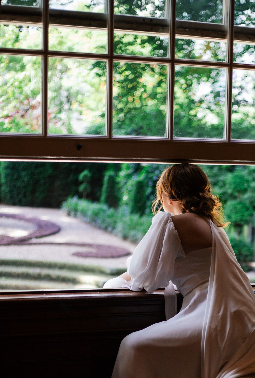 Bride in white gown gazing out the large window at a garden, elegant updo hairstyle, romantic wedding day moment in historic Amsterdam venue