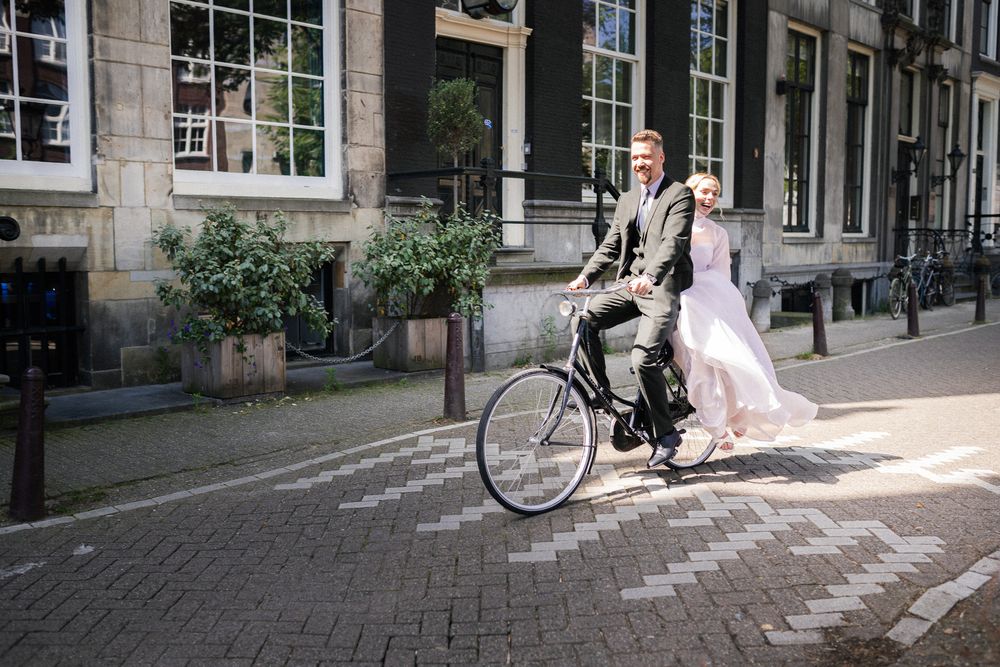 Bride and groom riding a bicycle together on cobblestone streets in Amsterdam city center on their wedding day.