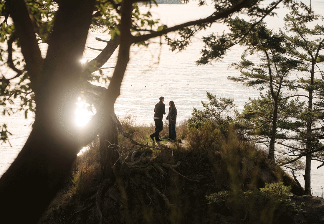 Two silhouetted figures stand on a hilltop at sunset among pine trees creating a dramatic backlit scene.