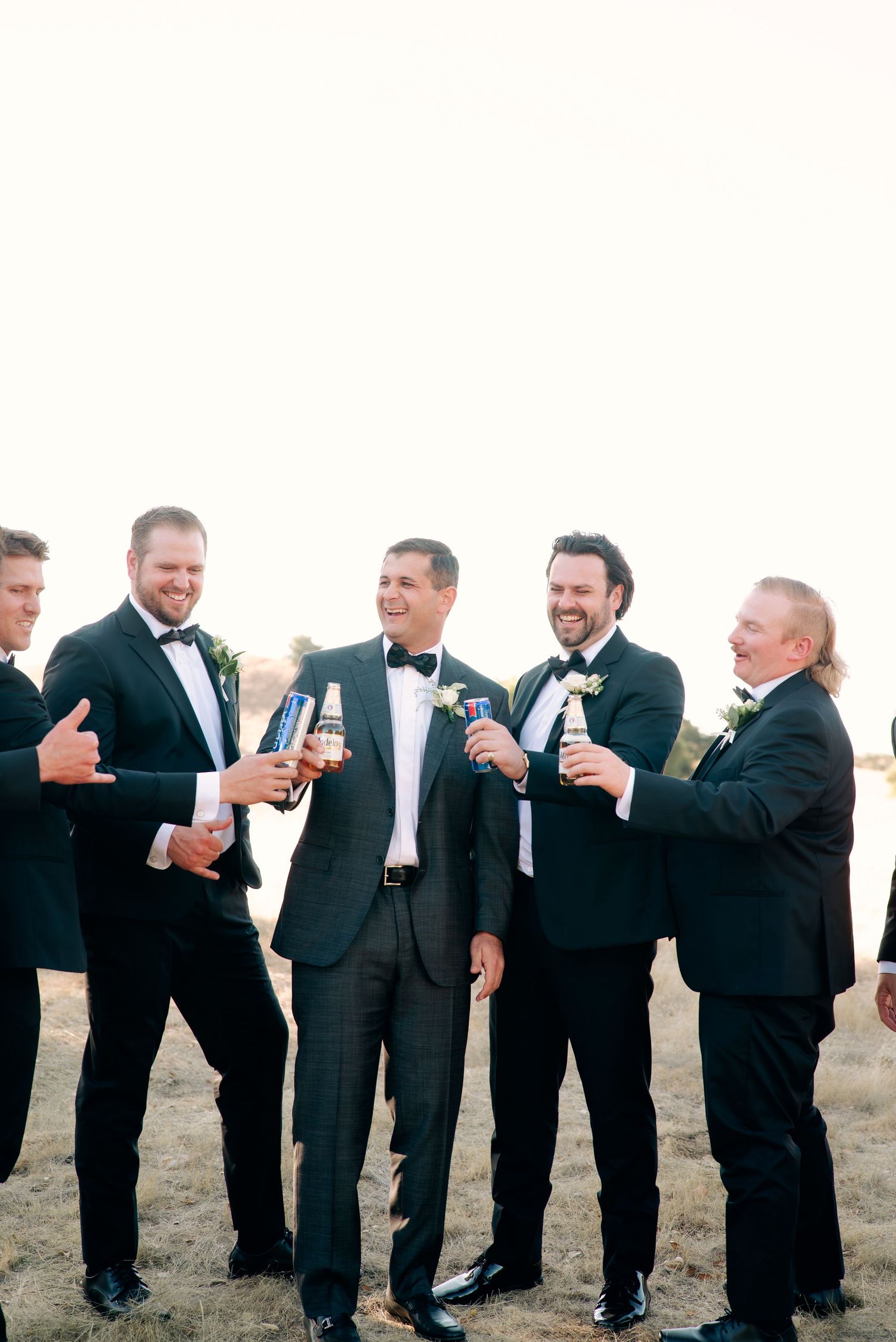 Groomsmen in black suits lined up outdoors toasting with drinks at a wedding celebration.