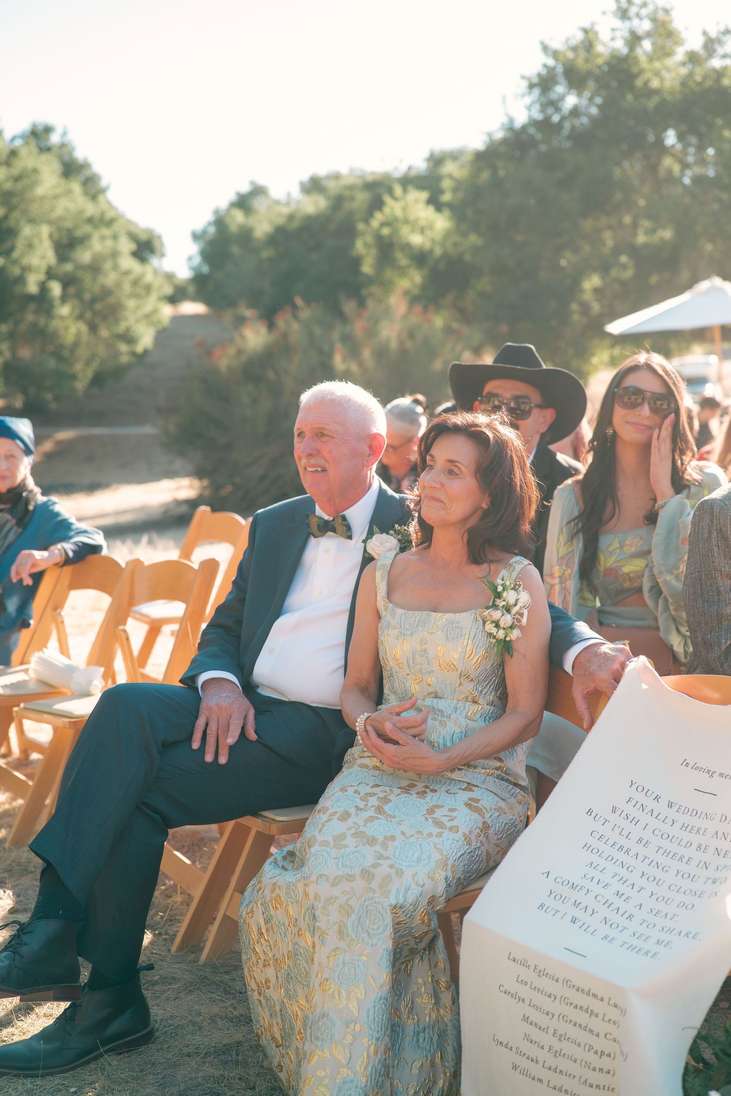 Guests seated in wooden chairs at an outdoor wedding ceremony with sunlit trees in the background.