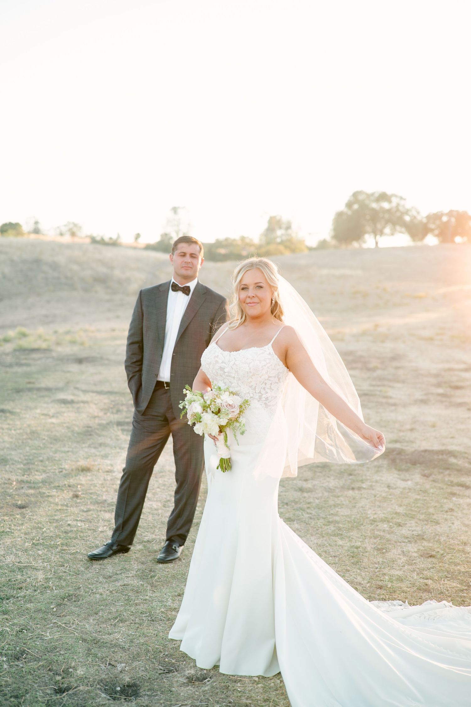 Bride in flowing white gown poses with her veil in the warm evening light.