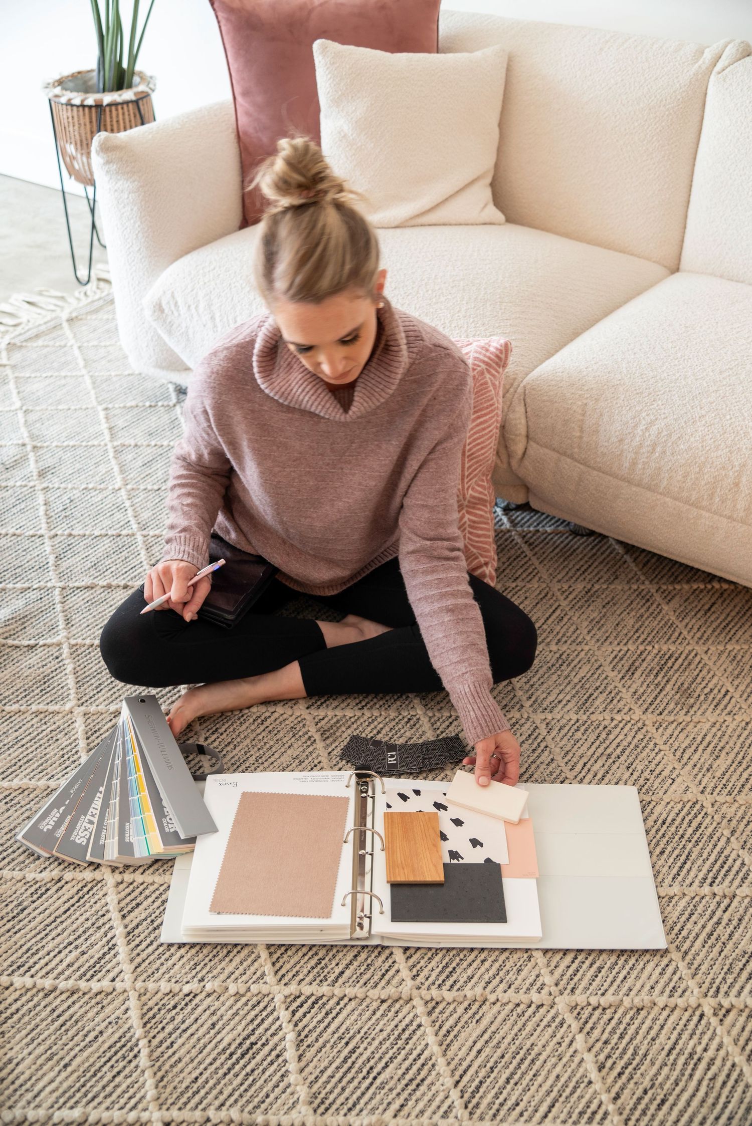 Someone in a cozy sweater sits cross-legged on a carpet reviewing design materials and color swatches by a white sofa.