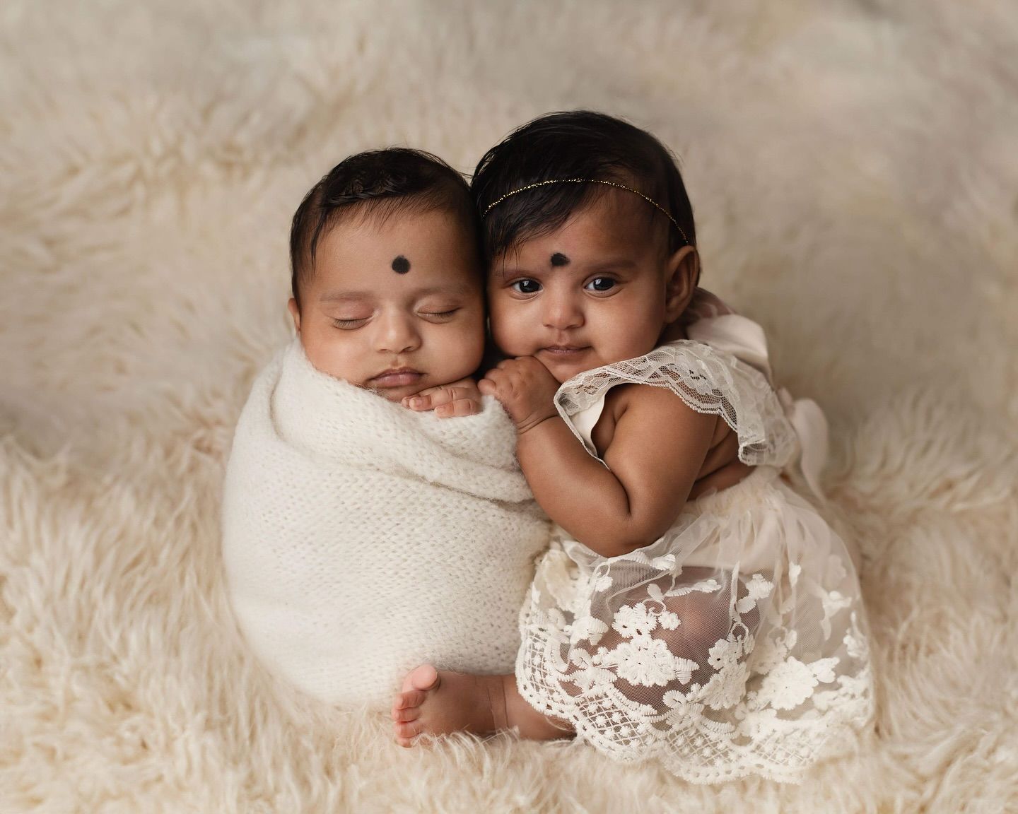 Two sleeping newborn babies in white swaddles and lace outfits lying close together on a soft cream backdrop. Elizabeth Lang Photography Sur