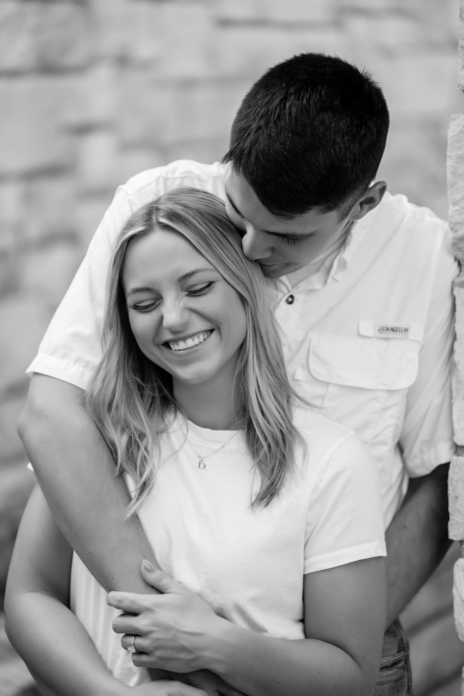 Black and white photo of a smiling blonde woman being embraced and kissed on the head by a young man in a white shirt.