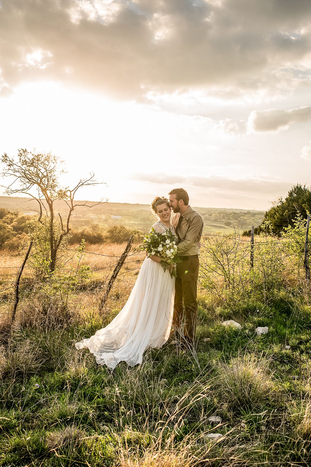 A couple in wedding attire embraces on a hilltop at sunset with a sweeping natural landscape in the background.