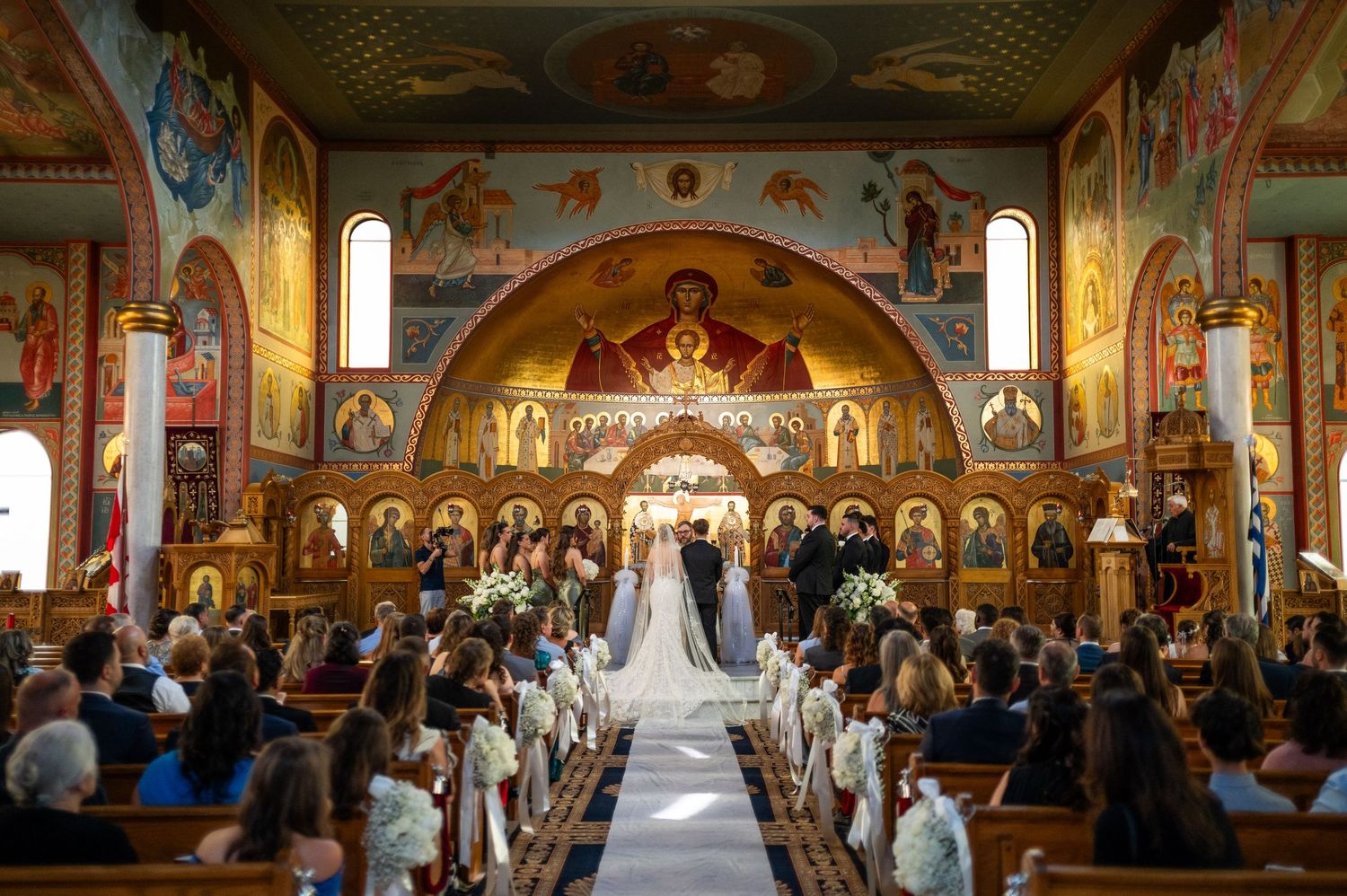 Orthodox wedding ceremony taking place in an ornate church with gilded icons and frescoes adorning the walls and ceiling.