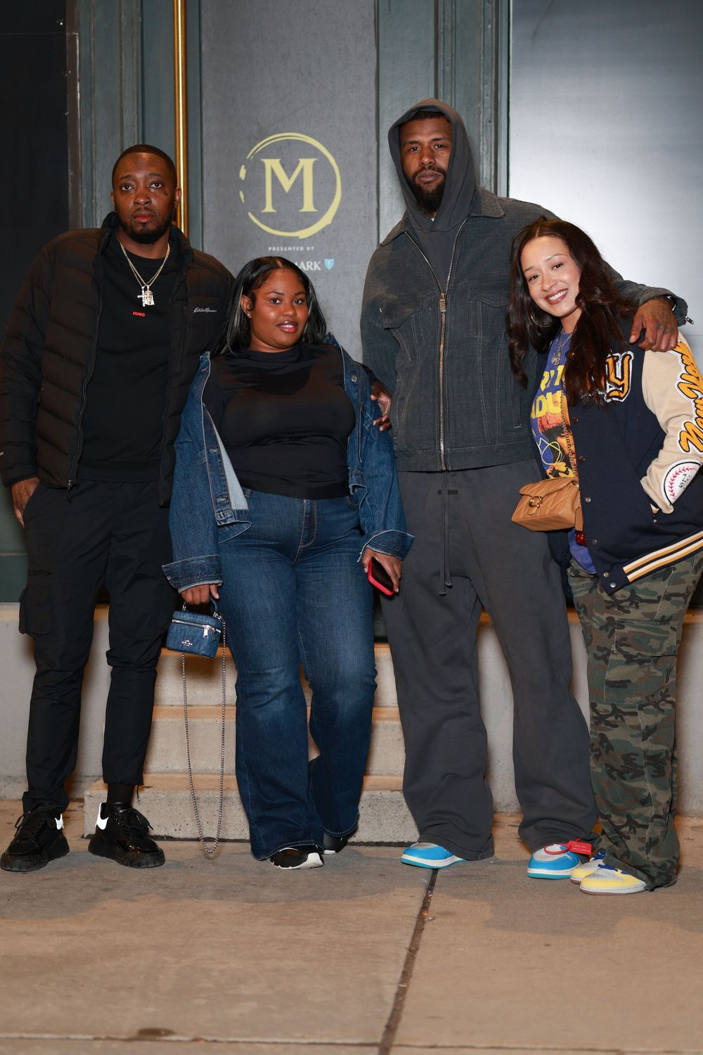 A group of four people posing together outside a building with an 'M' logo, wearing casual streetwear and varsity jackets.