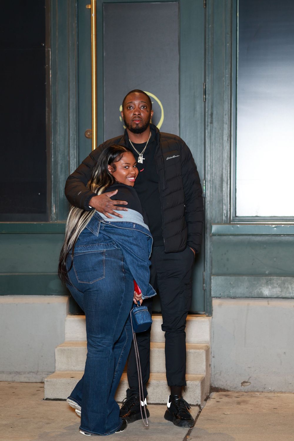 A couple in casual attire sits together on concrete steps outside a building with green walls and windows.