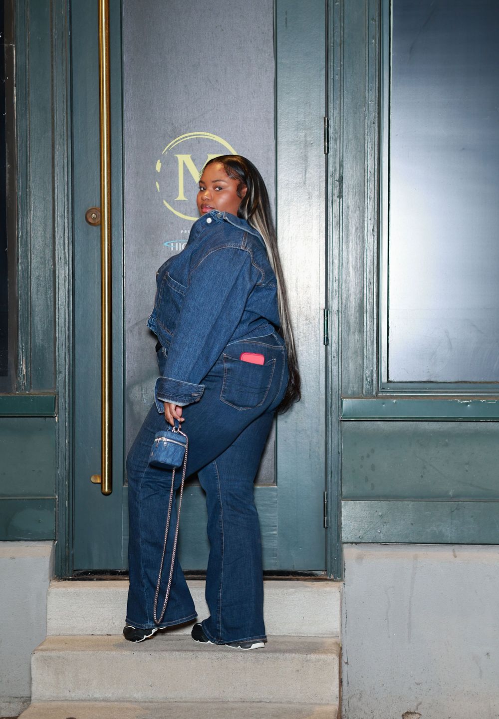 Person in blue denim workwear attempts to open a door with a gold handle in dim lighting.