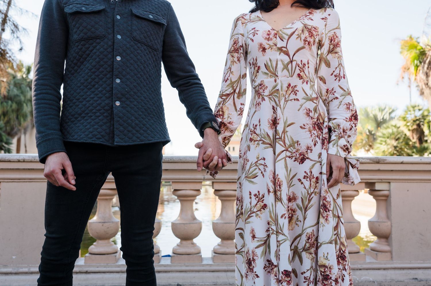 A couple holding hands outdoors, wearing a navy jacket and floral dress against a stone balustrade background.