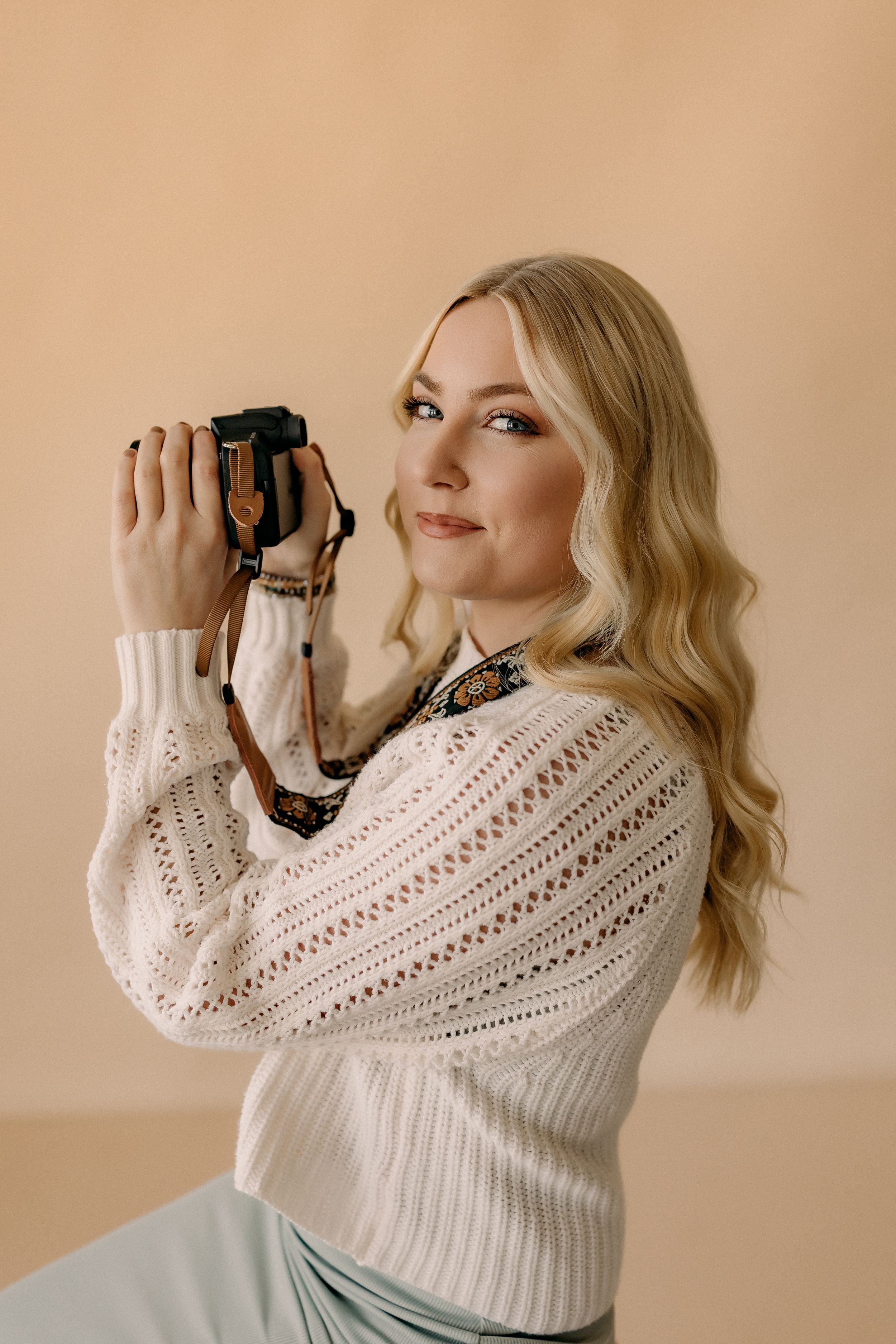 Photographer wearing white knit sweater holding vintage camera against beige background.