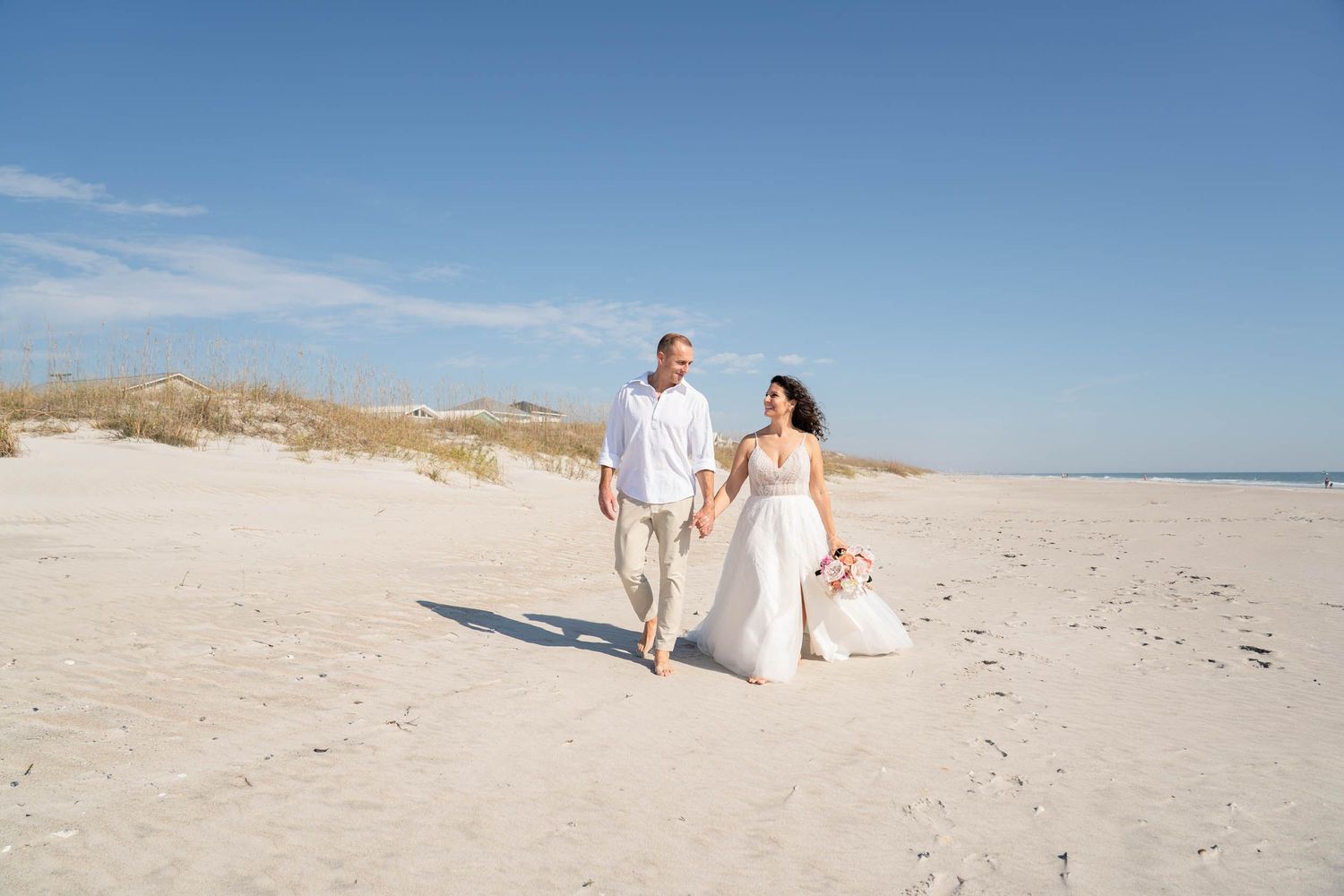 A couple strolls hand in hand on a sunny beach with sand dunes in the background.