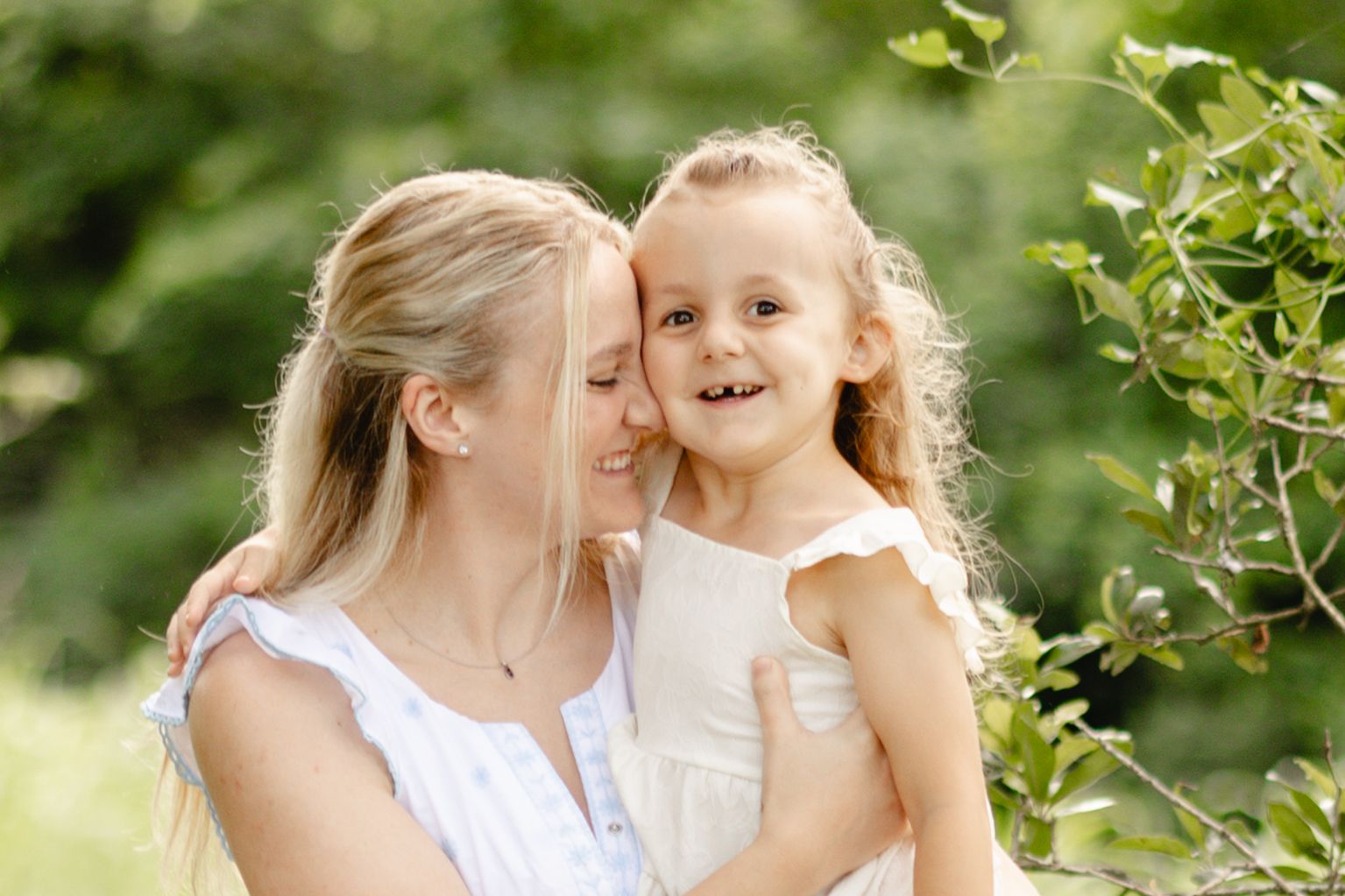 A smiling mother holds her happy young daughter outdoors, both wearing white dresses surrounded by lush green foliage.