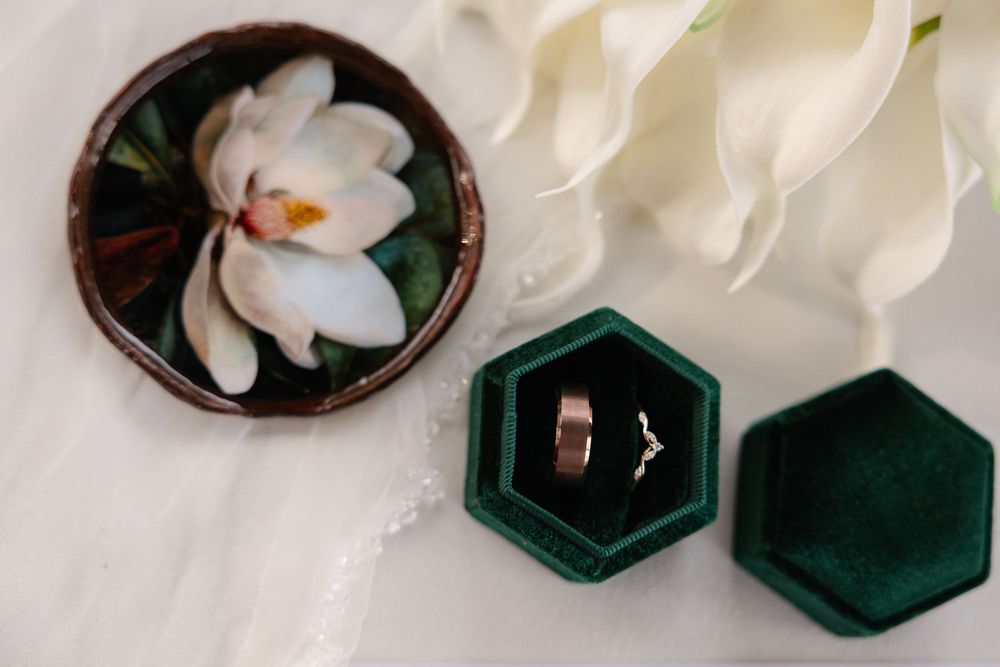 Wedding rings in dark green velvet hexagonal box next to white magnolia flower on light surface.